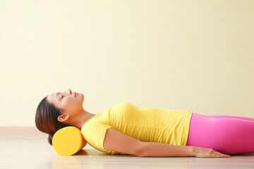 A woman lies on the floor using a yellow foam roller under her neck for muscle relaxation and stretching in a calm indoor setting.