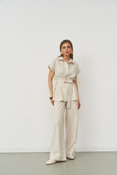 Woman in beige outfit stands confidently with hands at sides, wearing short-sleeved shirt, belted waist, wide-legged pants. Minimalist style with neutral colors. She exudes elegance and poise