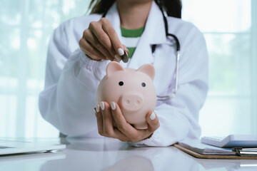 Close up young female general practitioner putting coin in piggy bank, managing hospital budget...