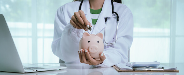 Close up young female general practitioner putting coin in piggy bank, managing hospital budget using laptop accounting applications, medical insurance finances, healthcare money savings concept.