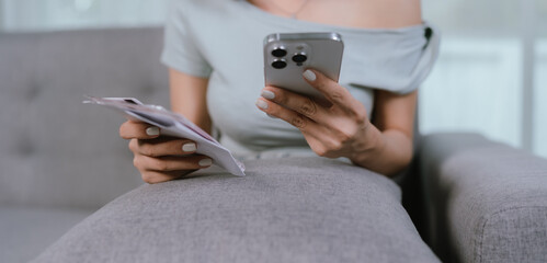 A woman checks her smartphone while holding paper bills, managing personal finances and expenses at home, with credit cards nearby on a couch in a relaxed setting.