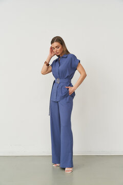 Woman wearing a blue jumpsuit with a belt, posing with one hand touching her hair and the other on her hip. She wears white sandals and a bracelet, standing in a minimal setting