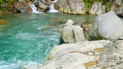 A refreshing waterway flows beneath a large cliff rock, framed by dense greenery and cascading...