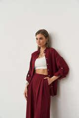 Woman in stylish burgundy outfit with a white crop top leaning against a white wall. She has long...