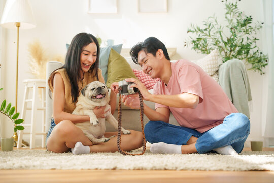 Asian man and woman enjoy playful moment photographing with camera with their adorable dog at home, sitting on rug and smiling.