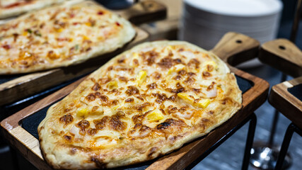 A freshly baked flatbread pizza topped with melted cheese and pineapple, served on a wooden tray in a restaurant or food market setting.