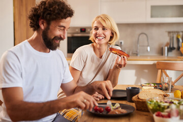 A couple is seated at a dining table in a homey kitchen, smiling and enjoying breakfast. The man is plating food while the woman holds a slice of toast, creating a warm atmosphere.