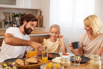 Family gathers in a bright kitchen to enjoy a hearty breakfast. The father pours orange juice while the daughter smiles. Warm atmosphere filled with love and tasty food creates a joyful experience.