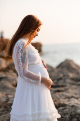 Smiling pregnant woman 25-29 year old in vintage white dress holding tummy standing over sea and...