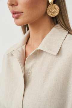Woman with smooth skin wears large round woven earrings and a cream-colored button-up shirt. Her lips are slightly parted, and the focus is on her lower face and shoulder area