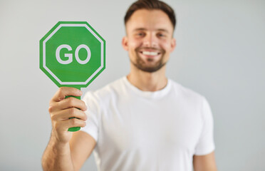 Portrait of young smiling man in white studio background, confidently holding green GO sign, as...