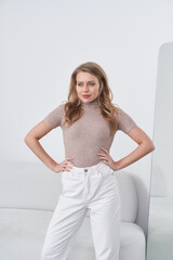 Confident woman stands with hands on hips, wearing a beige turtleneck and high-waisted white jeans....