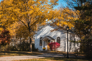 A white chapel  surrounded by vibrant fall foliage under a clear blue sky. The golden and red leaves create a warm, peaceful countryside scene perfect for seasonal and spiritual themes.