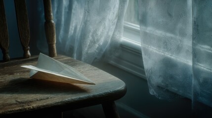A folded paper airplane rests on a wooden chair beside a window with sheer curtains