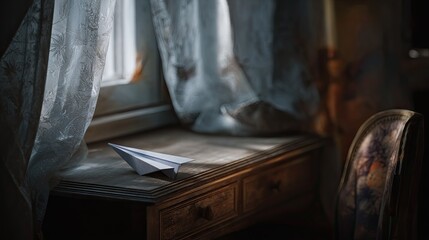 A paper airplane rests on a wooden desk by a window with sheer curtains. Soft light streams in