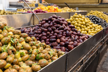 Colorful piles of marinated green, black, and purple olives arranged in metal trays at an open-air market. Fresh herbs, oil, and brine add texture and color to the authentic Mediterranean scene.