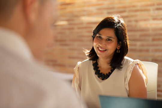 A young woman, with Indian Asian features, smiles while chatting with her teammates during a meeting in the office in the office