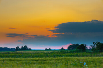 An evening landscape with a transition from a bright yellow sky to dark blue clouds over green fields and a tree-lined horizon. This image conveys the tranquility and beauty of twilight, a picturesque