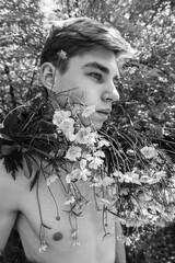 A shirtless young man poses outdoors with wildflowers draped over his shoulders
