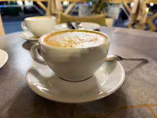 A white coffee cup with a white saucer sits on a table. A spoon is resting on the saucer
