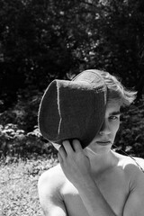 A shirtless young man covers one eye with a hat while holding a blade of grass in his mouth