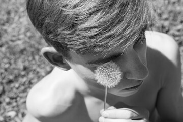 A shirtless young man holds a dandelion close to his eye while sitting outdoors