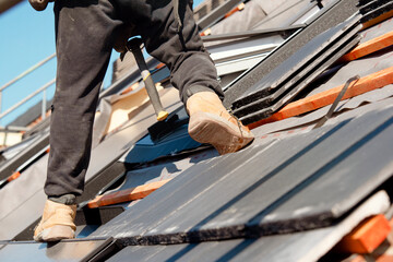 Roofer carefully places roof tiles on a sloped surface while standing on top of a building under a...