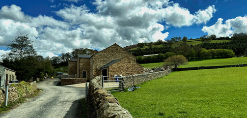 “Stone buildings line a country road beneath rolling green hills and a cloud strewn sky, with...