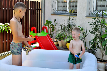 Two boys play in an inflatable pool surrounded by green plants on a warm summer day.