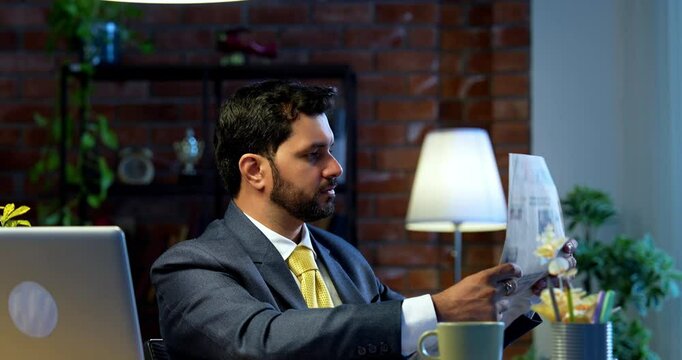 businessman reading business newspaper while sitting at modern computer desk, Indian Asian corporate executive focused on financial news, market updates, concept of awareness, leadership