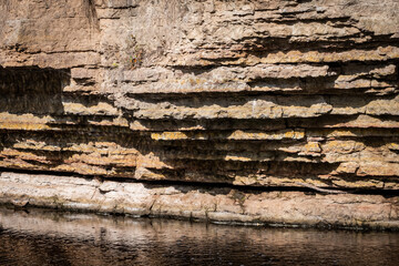 The yellow limestone wall of a canyon is cut by a stream flowing at its base. Sharp shadows...