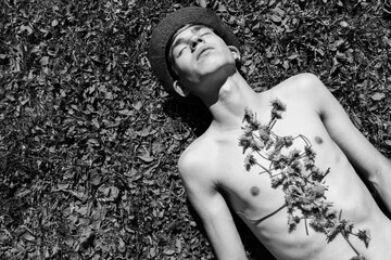 A shirtless young man in a hat lies on the grass with flowers arranged on his chest