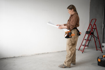 Repairwoman wearing tool belt holding paper blueprint while standing on construction site or inside...