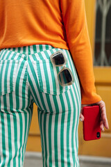 Woman in orange sweater and striped pants holding sunglasses and red smartphone.View from back view
