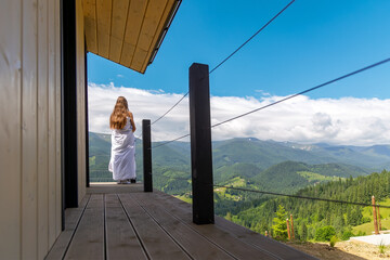 Woman on cabin balcony admiring mountain view. A woman wrapped in a white blanket stands on a...