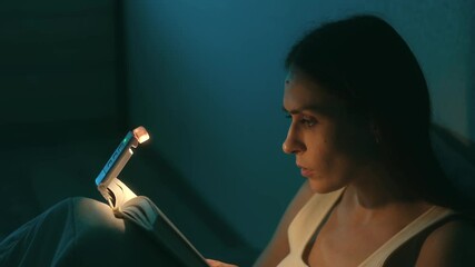Young woman reading book during nighttime, using small clip on led light illuminating page while enjoying quiet, peaceful moment of personal relaxation and literary immersion - Powered by Adobe