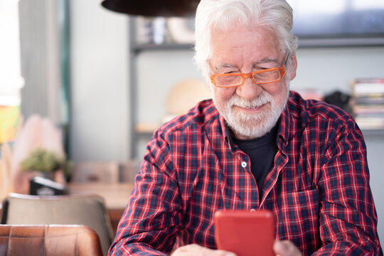 Smiling white-haired senior man sitting at cafe table using a smart phone with red cover. Elderly caucasian male in eyeglasses and checkered shirt