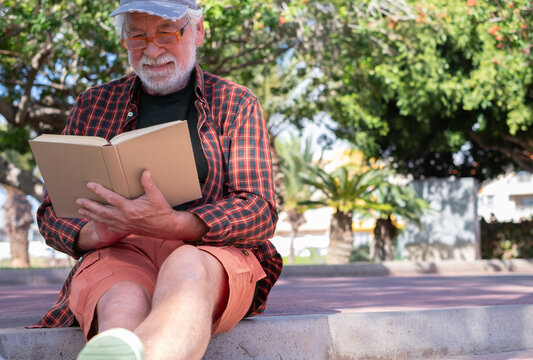 Serene retirement lifestyle. Smiling senior man sitting outdoors in public park reading a book. Bearded white-haired male with checkered shirt