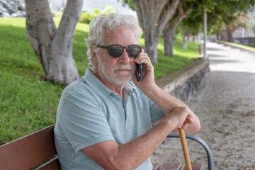 Serious white haired senior man with walking disability sitting in a bench in the park using mobile...