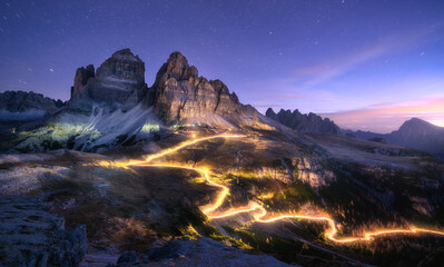 Car light trails on mountain road and high rocks at night in autumn in Tre cime, Dolomites, Italy. Colorful landscape with blurred light trails, hills, mountain peaks, sky with stars in fall at sunset