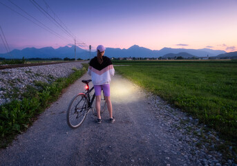 Woman is standing with mountain bike on gravel road at night in summer. Colorful landscape with sporty girl, bicycle with light, field, dirt road, green grass, purple sky at sunset. Sport and travel