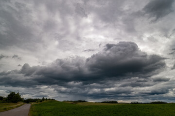 Dramatic storm clouds gather over a rural landscape with a narrow road winding through green...