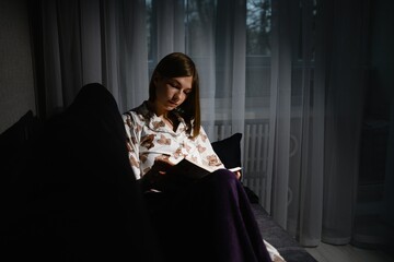 Young woman reading book in pajamas on sofa at night.