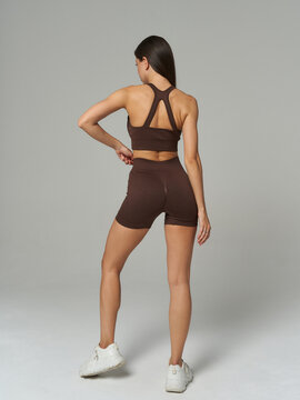 Athletic woman in brown workout outfit and white sneakers, standing with back to camera, right arm bent, left arm relaxed, on a neutral backdrop, long hair down the back