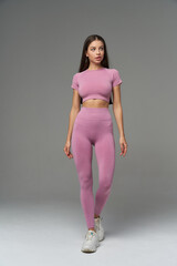 Woman with long hair in a matching pink crop top and leggings, wearing white sneakers. She stands...