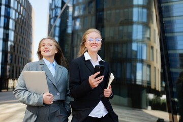 Two professional women walking confidently in a modern urban setting while engaged in conversation...