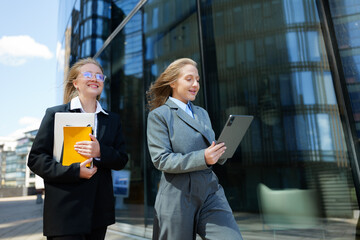 Two young professionals engaging with technology while walking outside a modern building on a sunny...