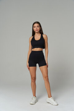 Athletic woman standing confidently in matching black sports bra and shorts, paired with white sneakers. Her long hair is down, and she maintains a neutral expression facing forward