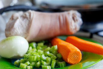 
A peeled pork shank is visible on a green surface, surrounded by fresh vegetables: sliced ​​celery stalks, half an onion, and two carrots.