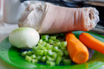 
A peeled pork shank is visible on a green plate, next to it is half an onion, chopped celery stalks, and two carrots.